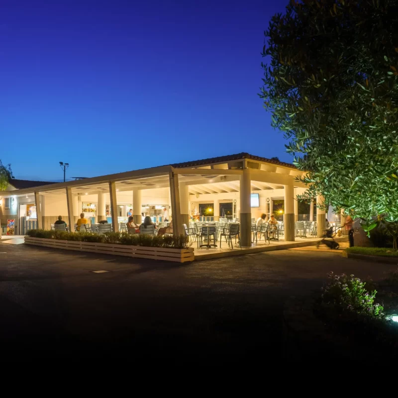 Locale all'aperto illuminato di sera con persone sedute ai tavoli sotto un cielo sereno.