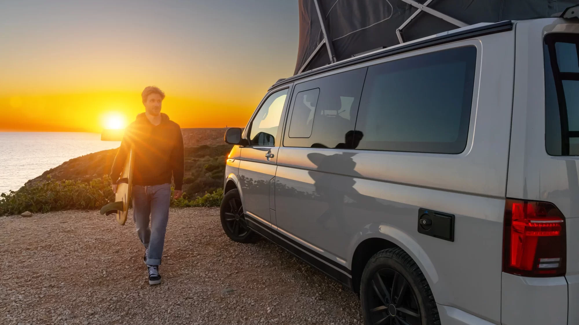 Un uomo con una tavola da surf cammina accanto a un camper al tramonto, accanto al mare.