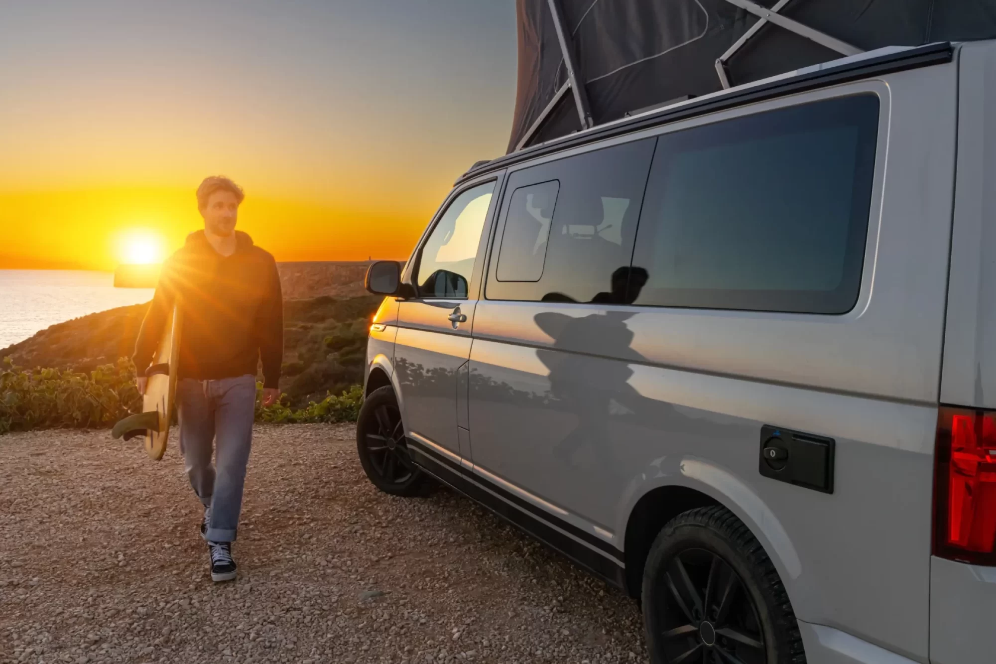 Un uomo con una tavola da surf cammina accanto a un camper al tramonto, accanto al mare.