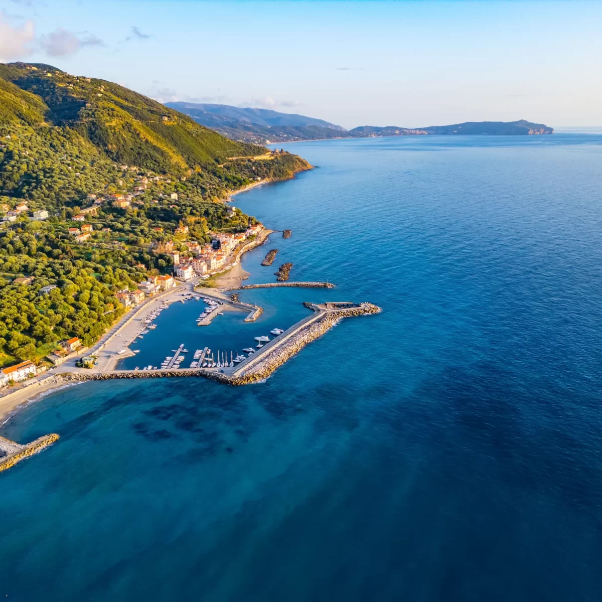 Veduta aerea di un porto costiero circondato da colline verdi e un mare blu brillante.