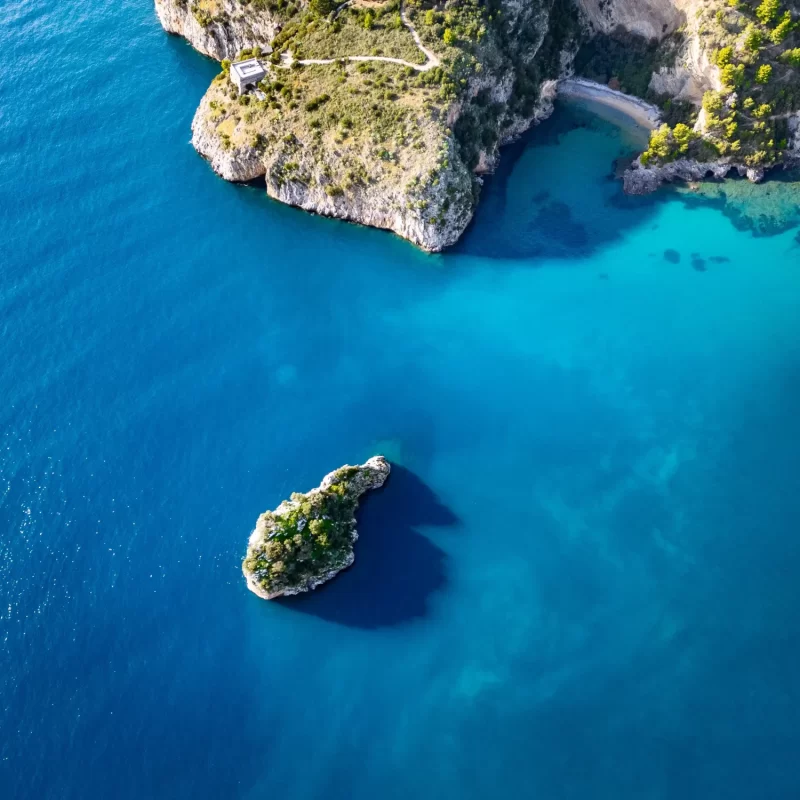Vista aerea di una costa rocciosa con acqua blu e un isolotto ricoperto di vegetazione.