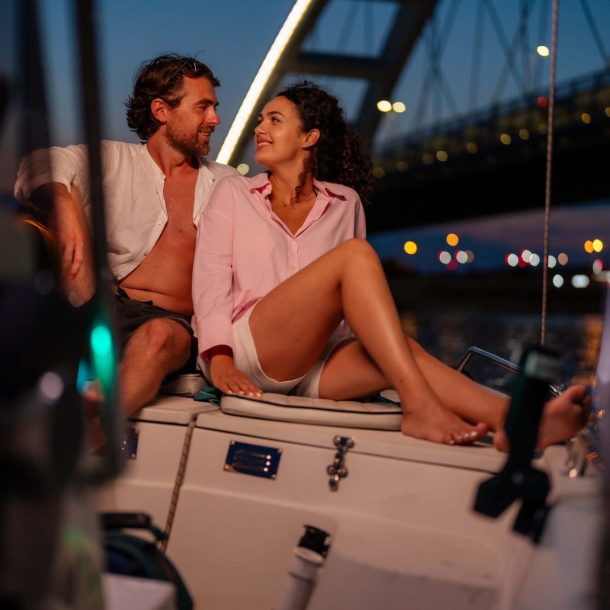 Coppia seduta su una barca al tramonto con un ponte illuminato sullo sfondo.