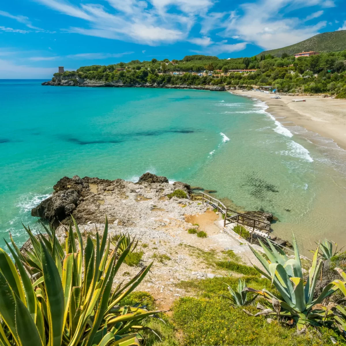 Spiaggia sabbiosa e costa rocciosa con mare turchese e vegetazione sotto un cielo azzurro.