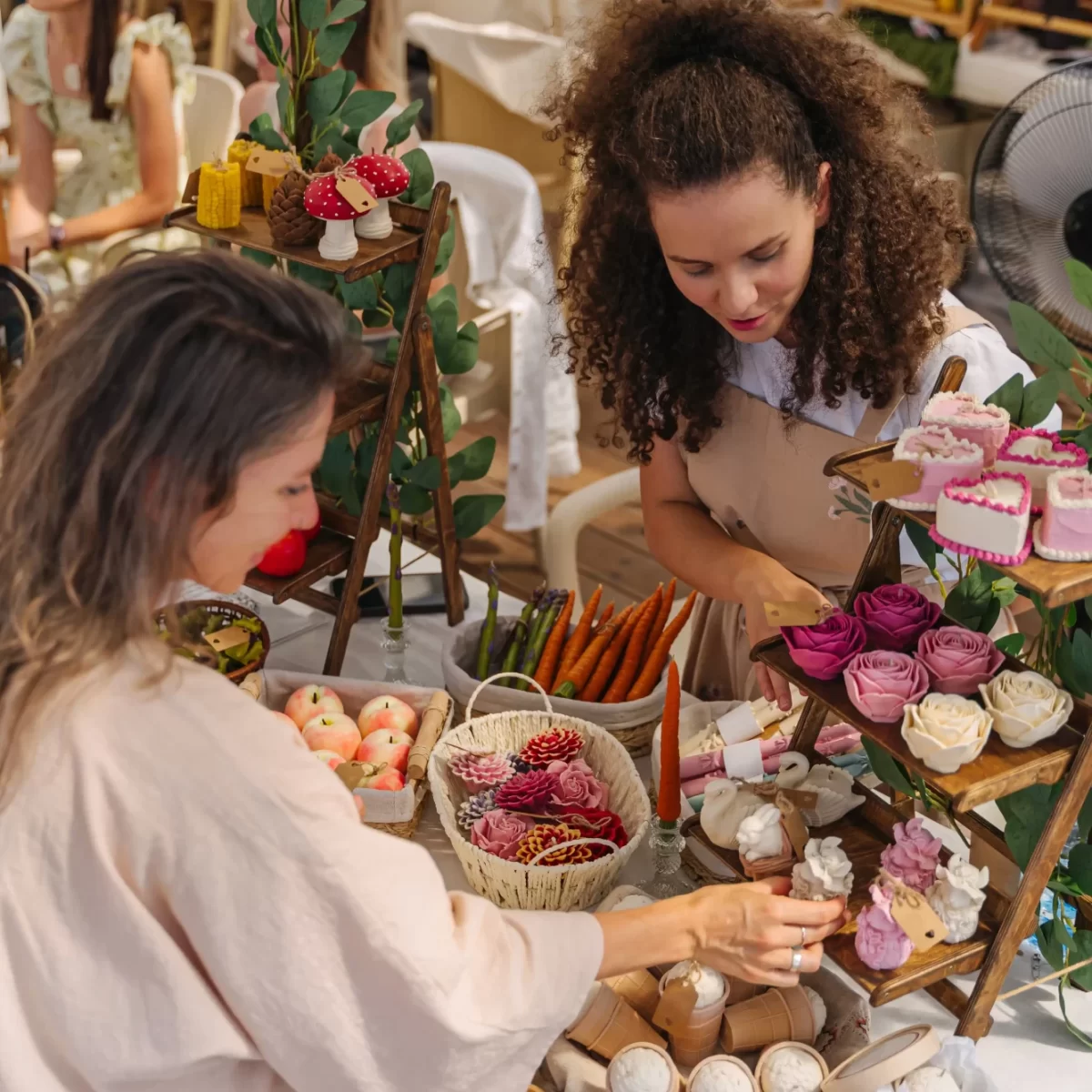 Due donne che scelgono candele artigianali a forma di fiori e dolci su un banco di mercato.