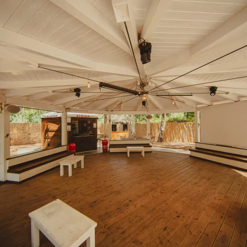 Area all'aperto con tetto in legno bianco, tavoli e sedute su pavimento in legno.