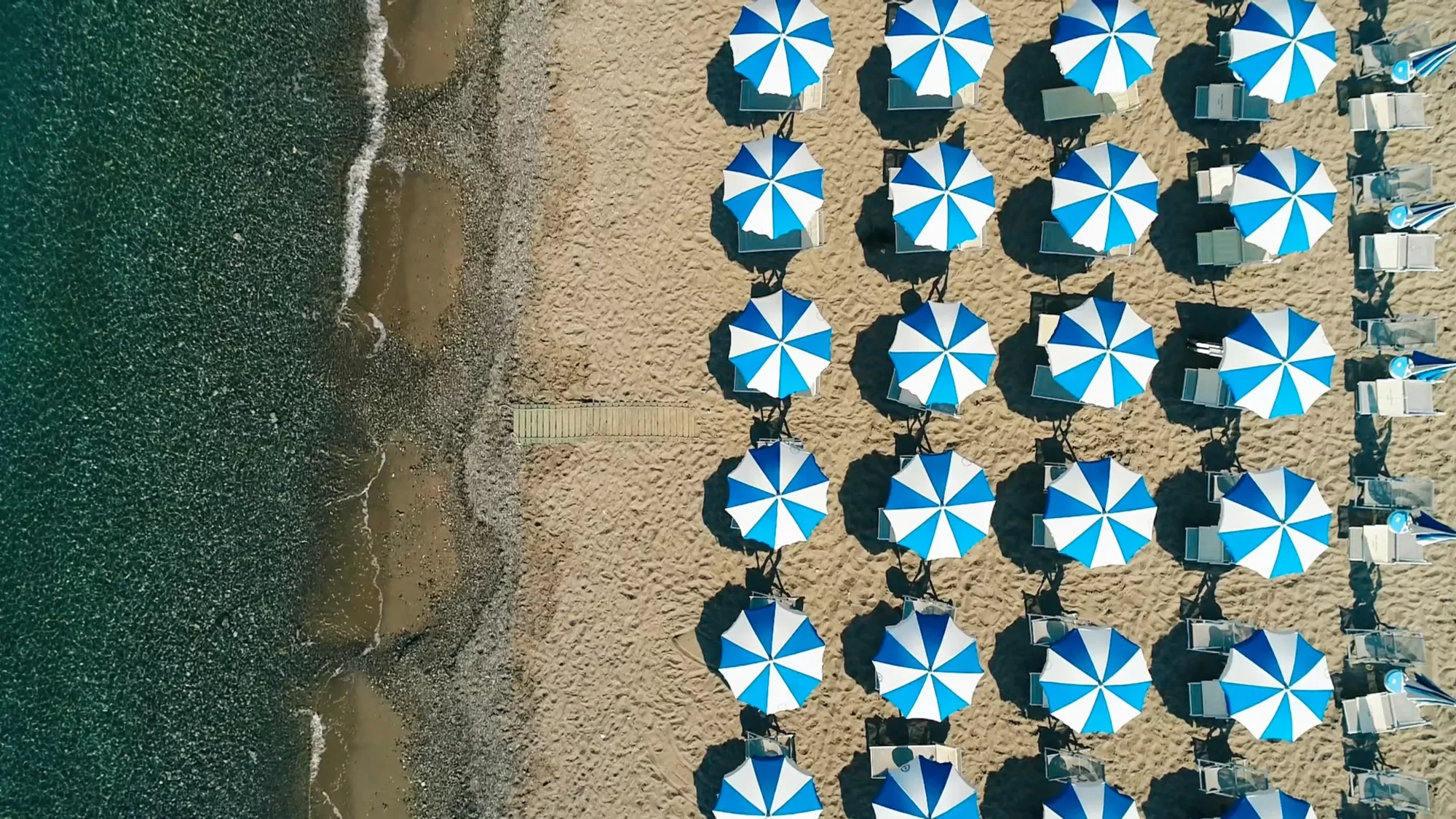 Spiaggia vista dall'alto con ombrelloni blu e bianchi disposti ordinatamente.