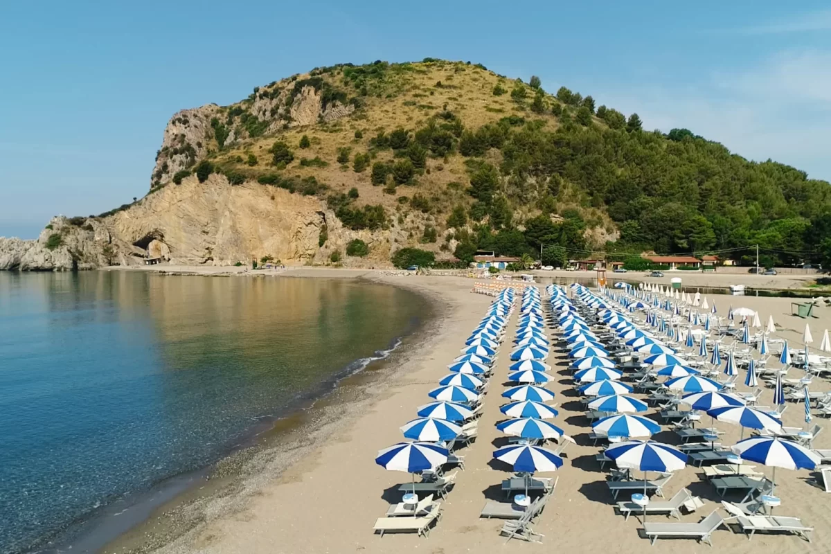 Spiaggia con file di ombrelloni blu e bianchi, montagne sullo sfondo e mare calmo.