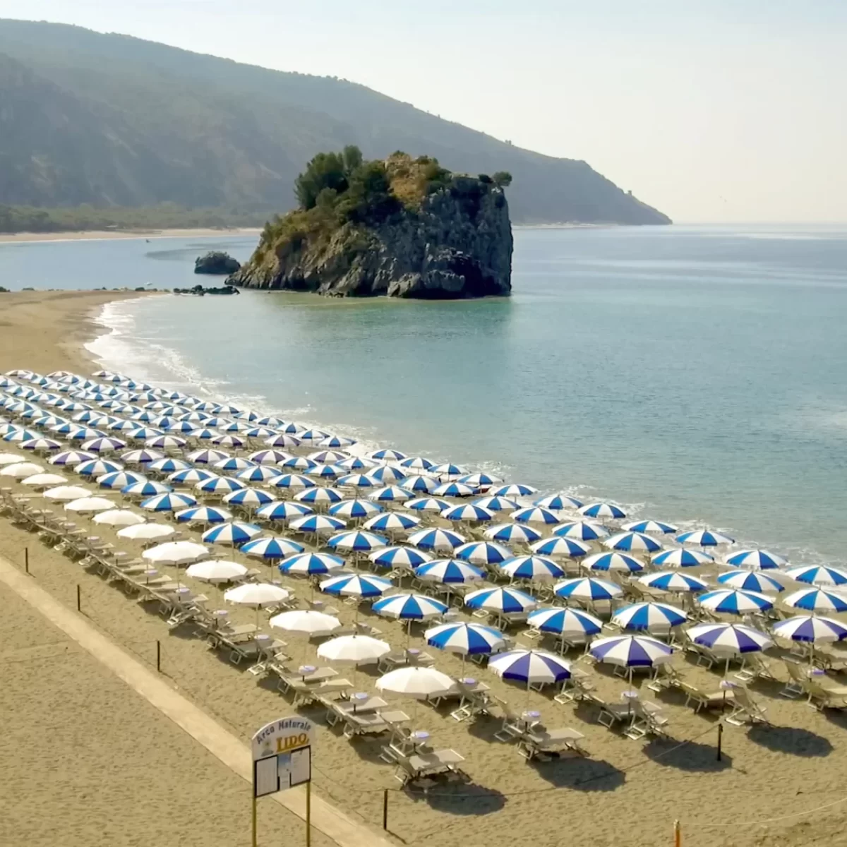 Spiaggia con file ordinate di ombrelloni blu e bianchi e un promontorio roccioso sullo sfondo.