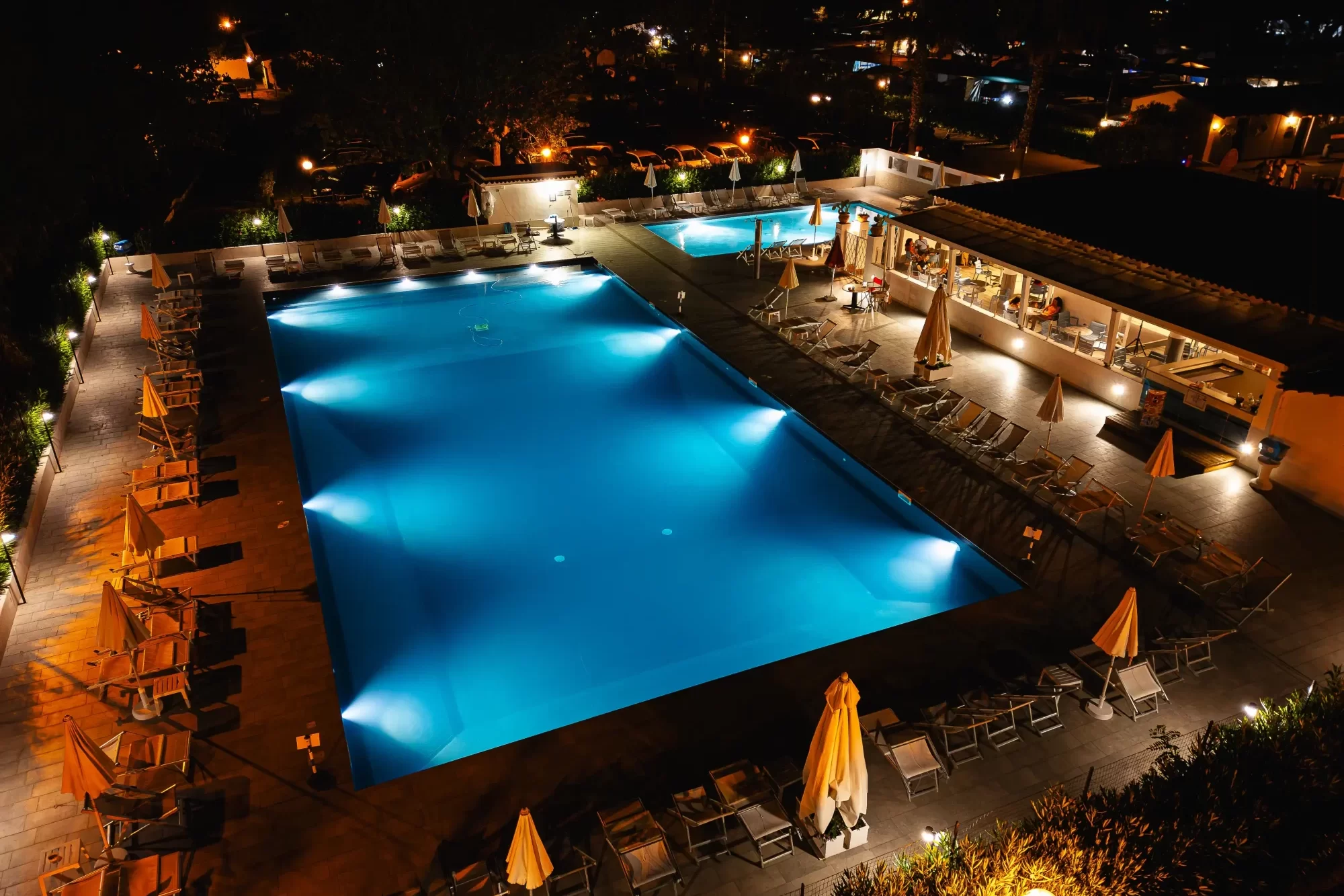 Piscina illuminata di notte con lettini e ombrelloni intorno.