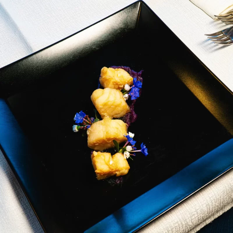 Cubetti di pesce impanati su piatto nero guarniti con fiori blu.