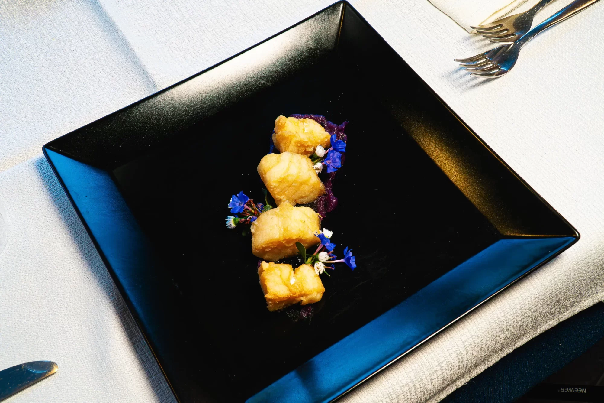Cubetti di pesce impanati su piatto nero guarniti con fiori blu.