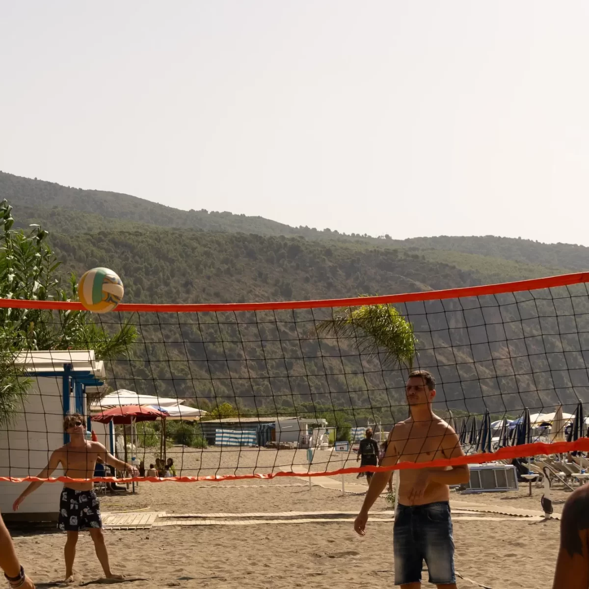 Persone che giocano a beach volley su una spiaggia con sfondo montuoso.