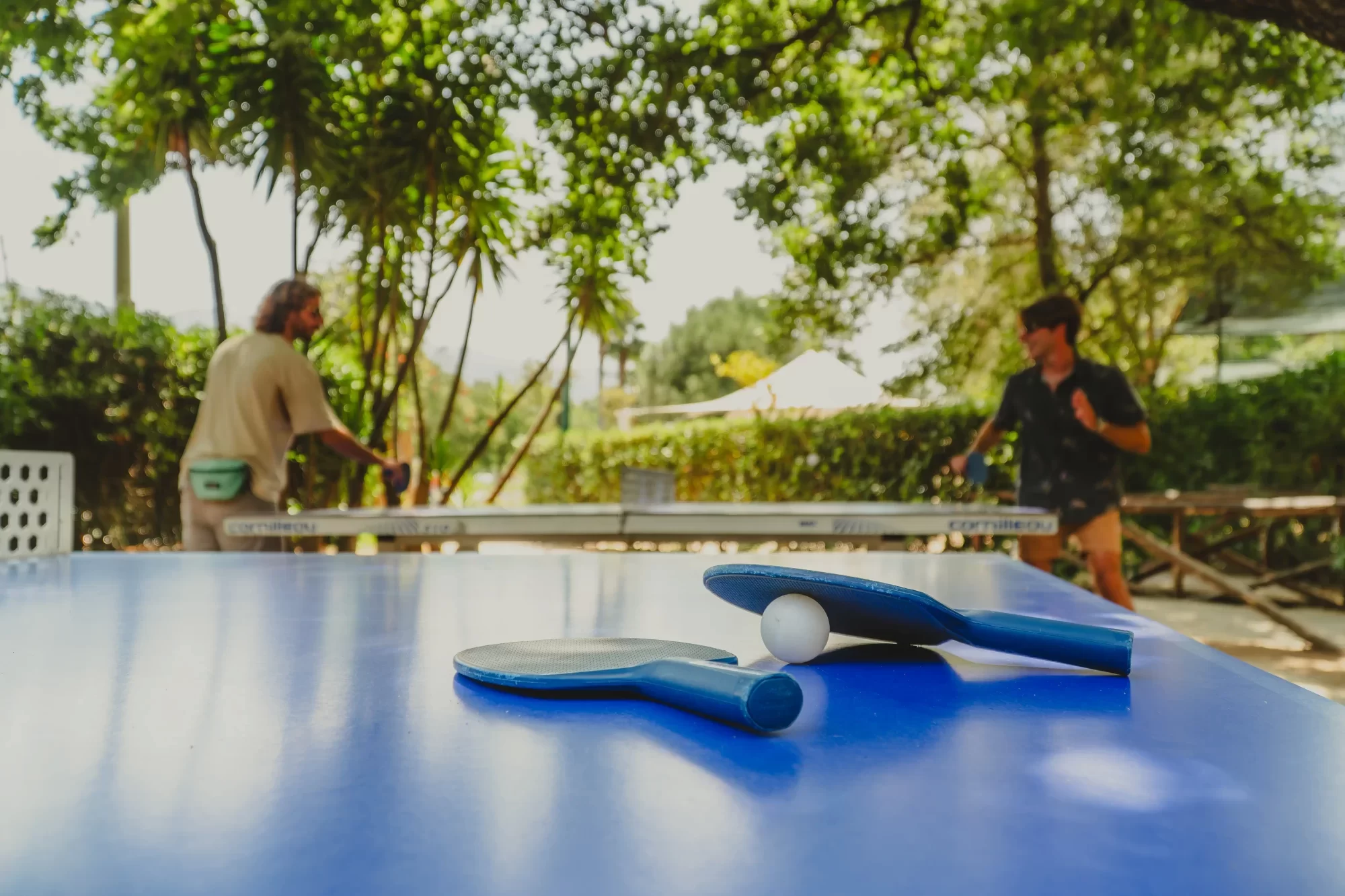 Due racchette da ping pong e una pallina su un tavolo blu con persone che giocano sullo sfondo.