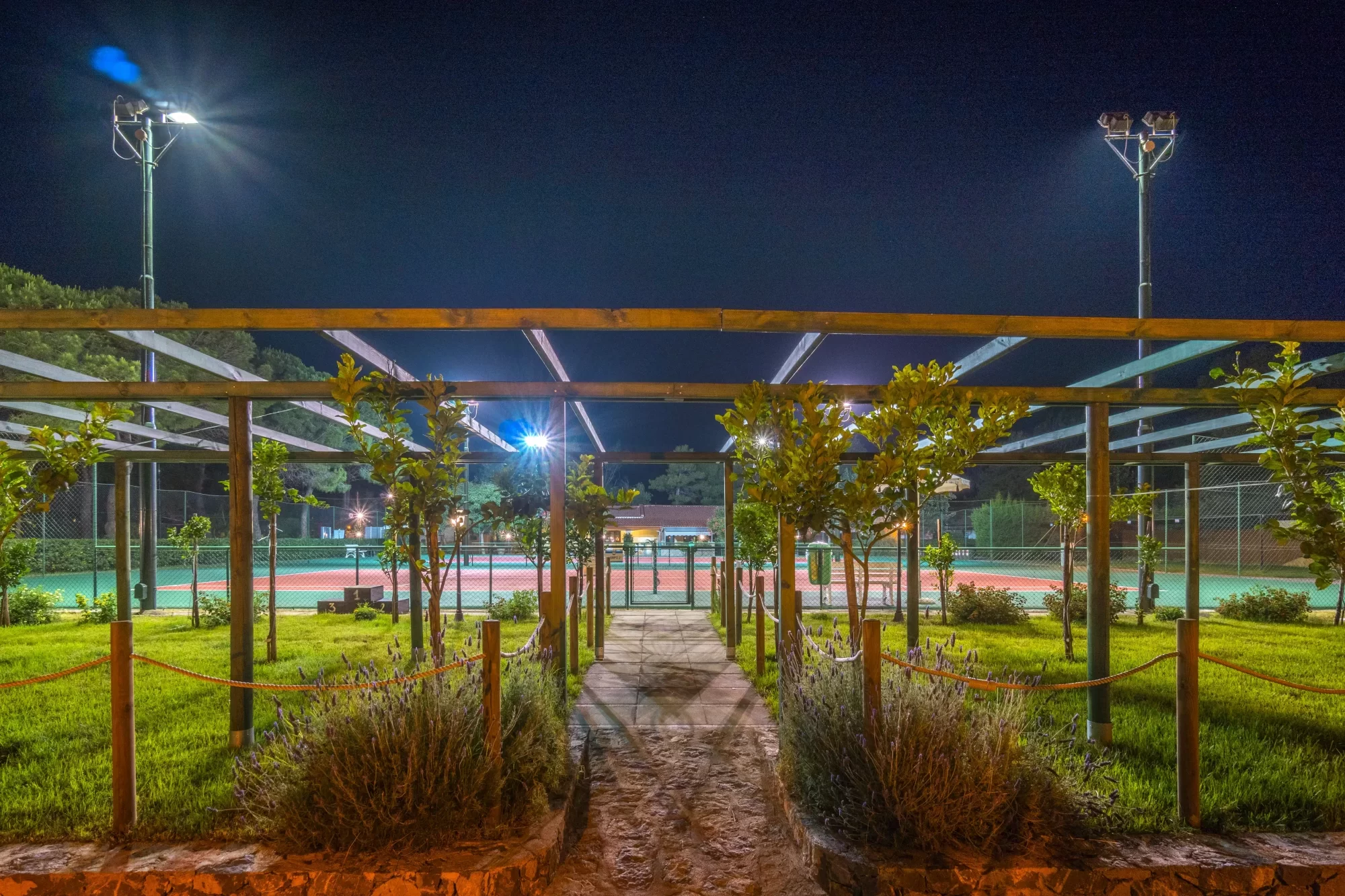 Percorso illuminato di notte che conduce a un campo da tennis circondato da giardini.
